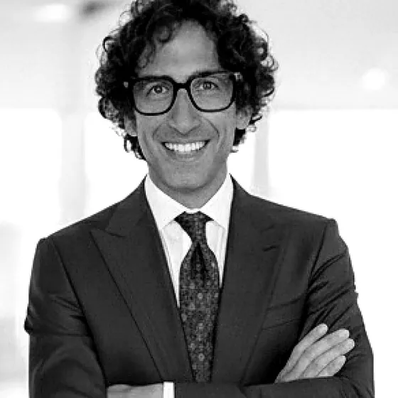 Ritratto in bianco e nero di Donato Marangelo, co-fondatore di MEMO: uomo sorridente con capelli ricci scuri, occhiali dalla montatura nera, giacca e cravatta, braccia conserte